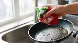 The strawberry scrubber covered in suds, cleaning a dark frying pan under running water